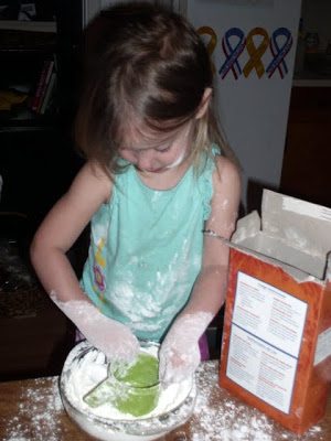 A young girl with flour on her hands and face is concentrating as she makes a mess in the kitchen. She is scooping flour from a white bowl with a green measuring cup. A cereal box is visible on the table. Ribbons hang on a wall in the background.