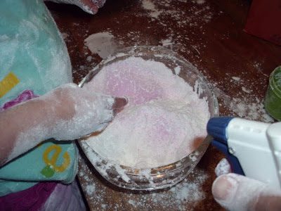 A child with flour-covered hands mixes a bowl of flour on a table. A spray bottle is partially visible, held by an adult. The table and the surrounding area are dusted with flour, creating a messy, playful scene.