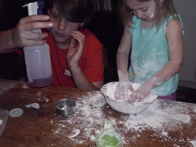 A child and an adult are making a mess with flour in the kitchen. The child is mixing flour in a bowl, while the adult holds a spray bottle. Flour is scattered on the table, along with measuring cups.