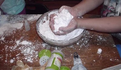 Two hands can be seen mixing a white powdery substance in a large metal bowl. The table is dusty with the same substance, and kitchen utensils and containers are scattered around.