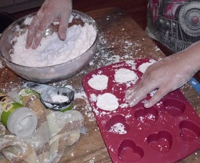 Hands press white powdery mixture into red silicone molds shaped like circles and hearts on a wooden table. A bowl with more of the mixture and kitchen items are visible, with a messy dusting of the mixture on the surface.