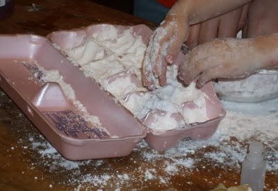 Hands playing with flour in pink egg carton, making a mess on a wooden surface.