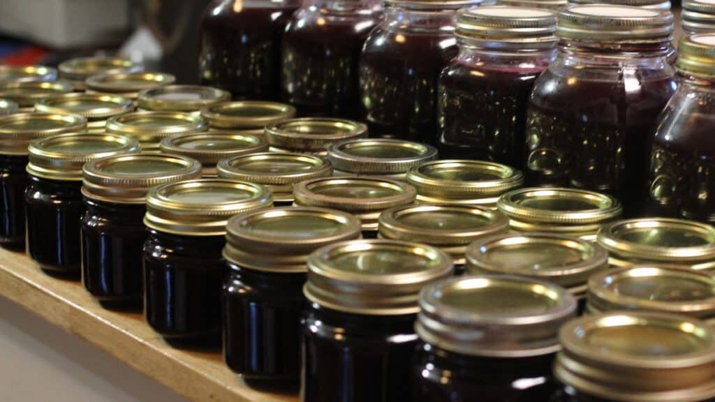 Rows of glass jars filled with dark purple mustang grape jelly, each sealed with a metal lid, are arranged neatly on a wooden surface.