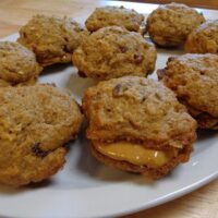 A plate of homemade oatmeal cookies, including one sandwich cookie filled with peanut butter. The cookies are golden-brown with visible oats and raisins. They are arranged on a white plate on a wooden table.