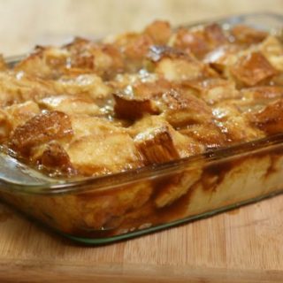 A glass baking dish filled with golden-brown baked french toast sits on a wooden surface. The bread cubes are baked and glazed, boasting a caramelized, slightly crispy top with a moist interior.