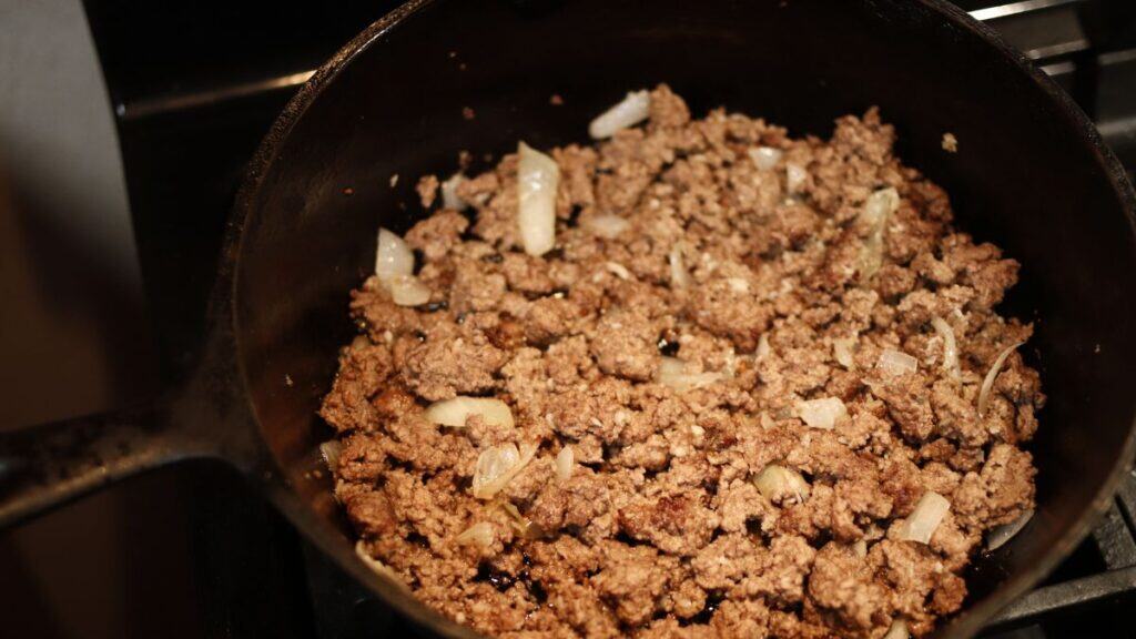 Ground beef and chopped onions cooking in a black skillet on the stovetop, forming the savory base for a classic shepherd's pie. The beef is browned and the onions are soft and translucent, ready for the next delicious steps.
