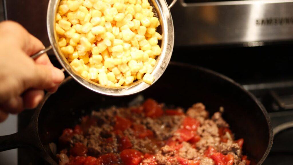 A hand holds a small strainer of yellow corn kernels above a skillet containing browned ground beef and diced tomatoes, preparing to add the corn to this shepherd's pie filling on the stovetop.