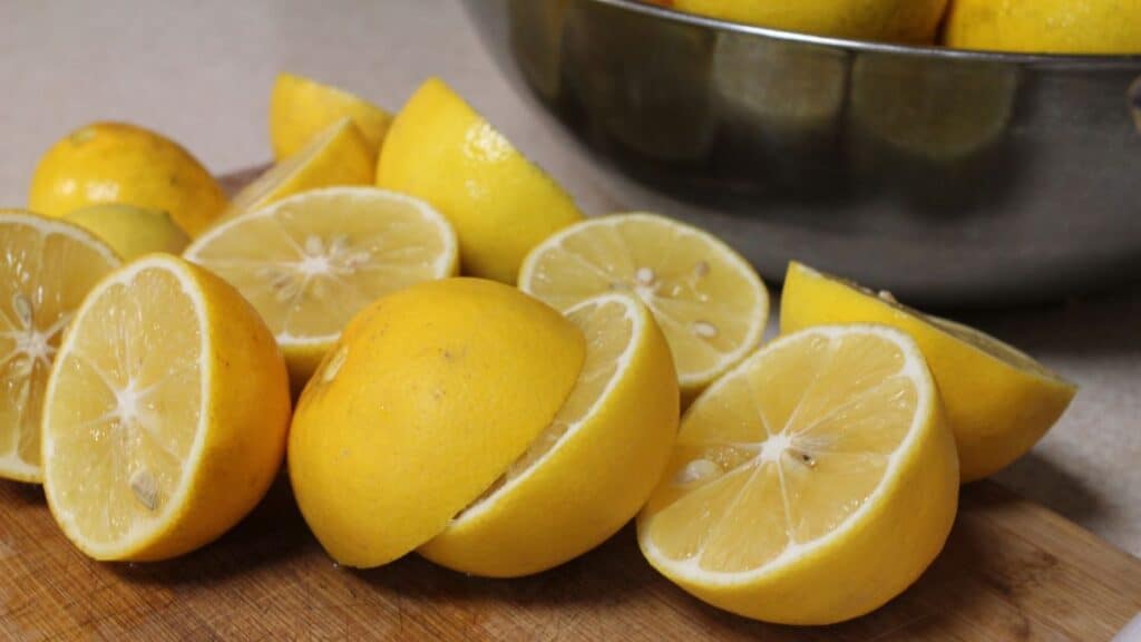 Several lemons are cut in half on a wooden cutting board, with more whole lemons in a large metal bowl in the background. The lemons are bright yellow and fresh, perfect for freezing lemons to preserve their vibrant flavor for later use.