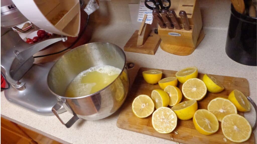 A metal bowl with lemon juice, freshly squeezed from frozen lemons, sits beside a cutting board with halved lemons on a kitchen counter. A stand mixer, knife block, and utensils are also visible.