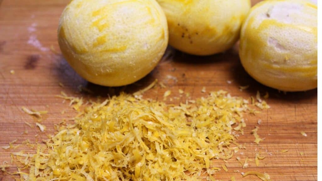 Three peeled lemons rest on a wooden cutting board with a pile of freshly grated lemon zest in front of them, perfect for freezing lemons or adding flavor to recipes with frozen lemons.