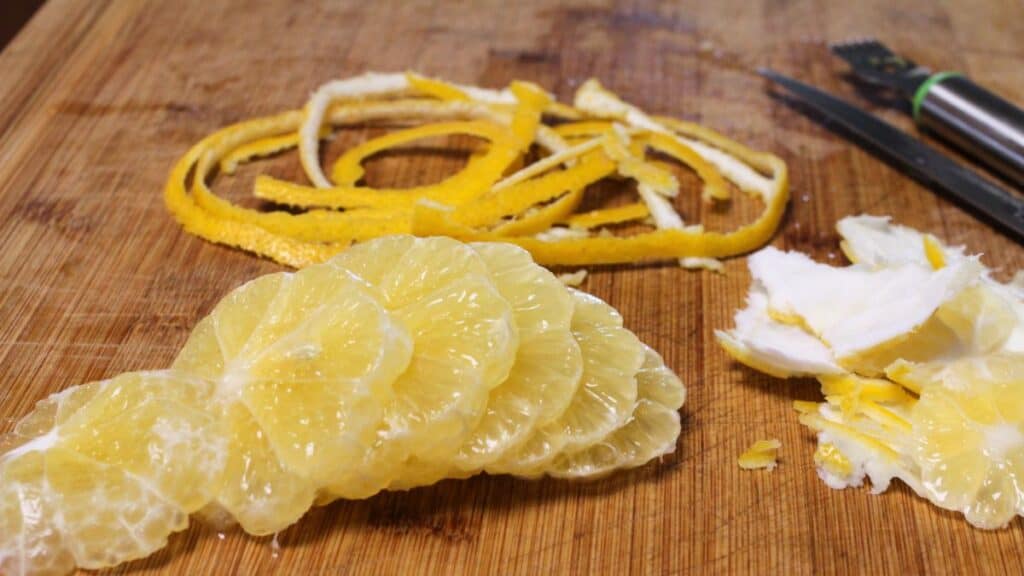 Sliced citrus fruit with yellow peel strips, white pith, and a zester on a wooden cutting board—ideal for preparing and freezing lemons to have fresh flavor on hand anytime.
