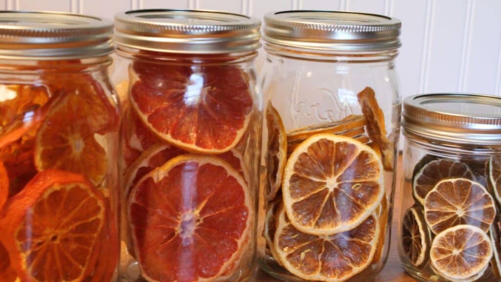 Four glass jars filled with dried citrus fruit slices, including dried lemon, oranges, and grapefruits, are lined up against a light-colored background.