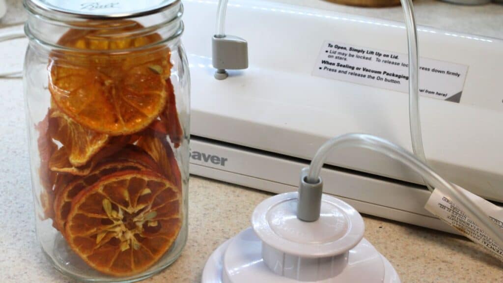 A glass jar filled with dried orange and dried lemon slices sits on a counter next to a white vacuum sealer machine with an attached hose and lid accessory.