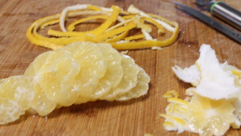 Sliced citrus fruit with yellow peel, white pith, zest strips, and dried lemon on a wooden cutting board, next to a metal peeler.