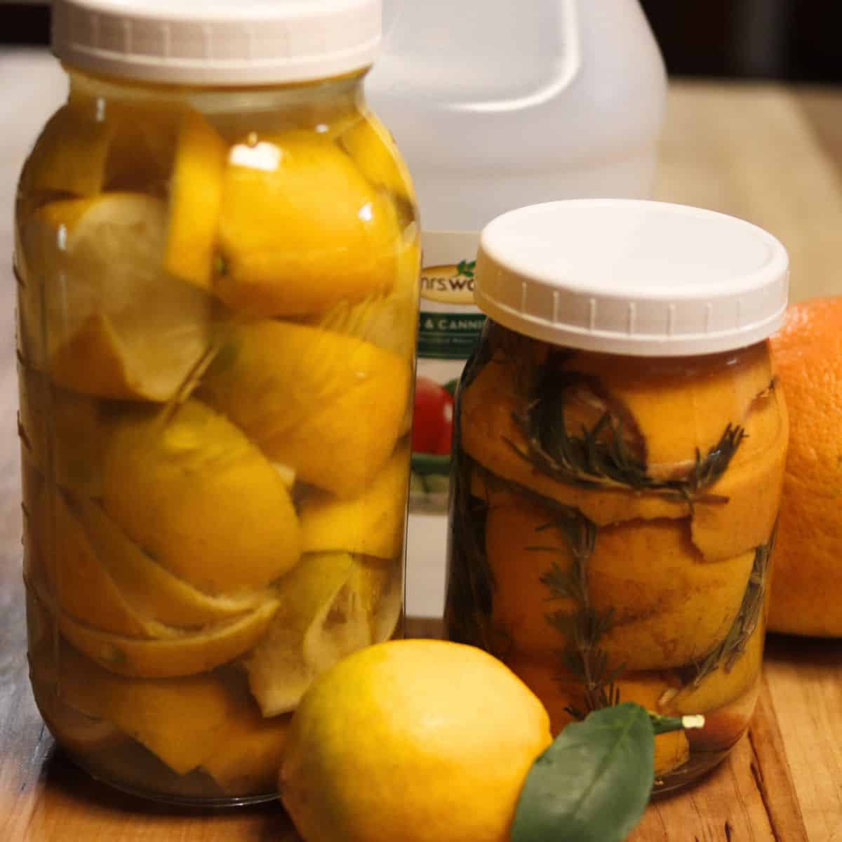 Two glass jars filled with preserved lemon slices and herbs sit on a wooden surface. A whole lemon with leaves, an orange, and a bottle of vinegar are in the background.