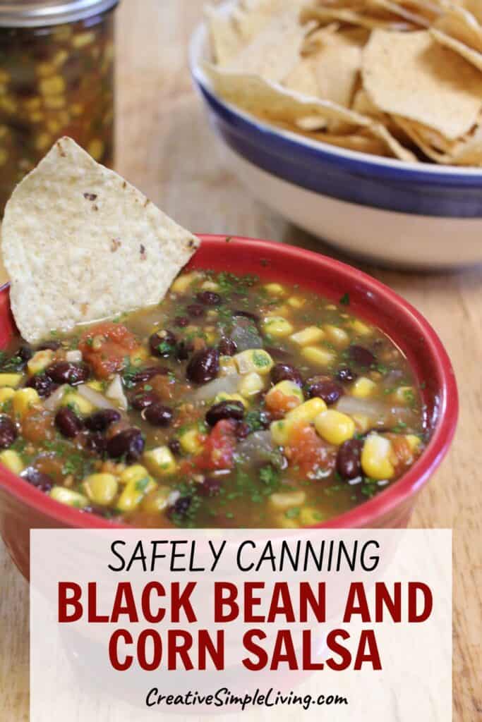A red bowl of canning black bean and corn salsa with a tortilla chip in it sits on a table. A bowl of chips and a jar of salsa are in the background. Text reads: "Safely Canning Black Bean and Corn Salsa.