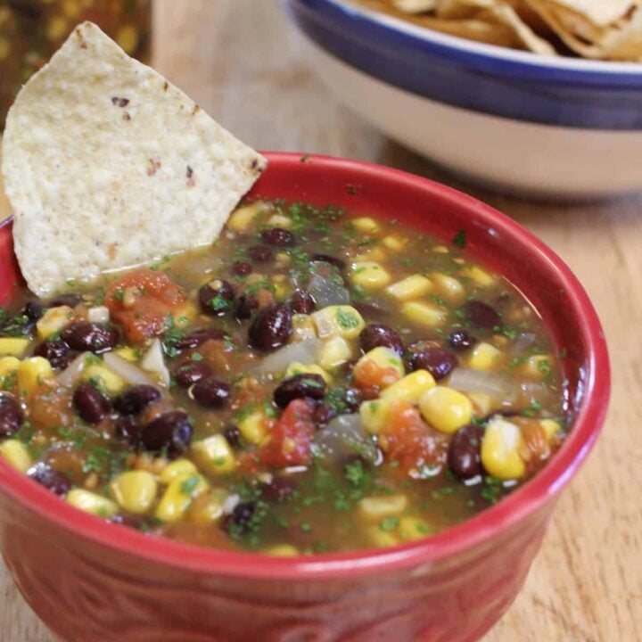 A red bowl filled with canning black bean and corn salsa, garnished with chopped herbs, sits on a wooden table. A tortilla chip is dipped in the salsa, and a bowl of more chips is visible in the background.