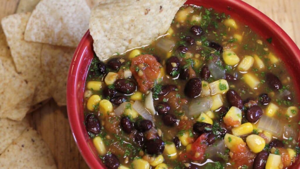 A red bowl filled with canning black bean and corn salsa, featuring tomatoes, onions, and herbs, with a tortilla chip dipped in the salsa. Extra tortilla chips are next to the bowl on a wooden surface.