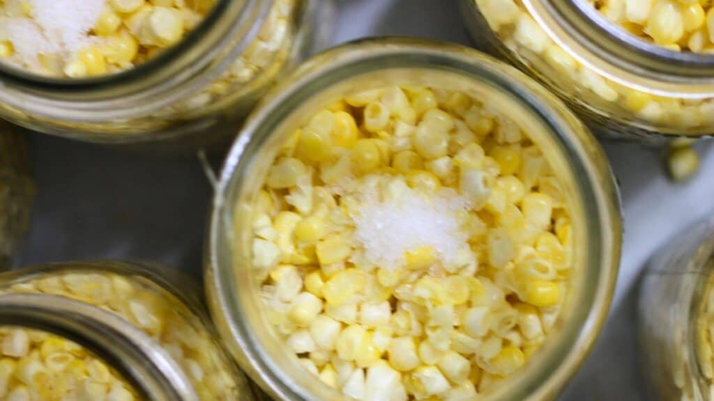 Overhead view of glass jars filled with yellow corn kernels, each topped with a small mound of white granulated salt, ready for canning corn at home.