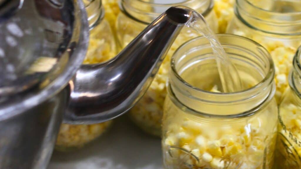 A metal kettle pours hot water into a glass jar filled with corn kernels, demonstrating the process of canning corn, with several similar jars in the background.