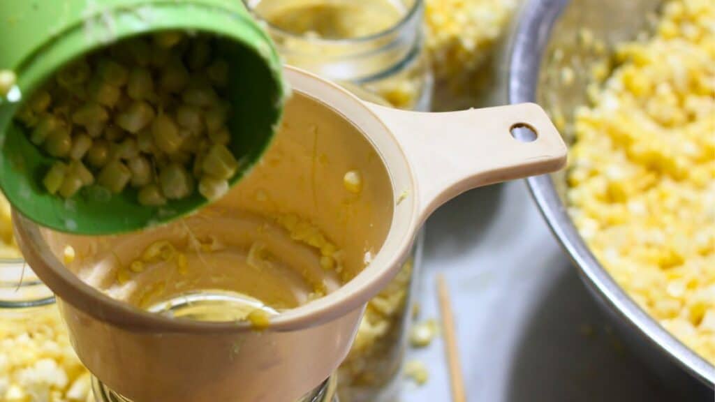Close-up of canning corn as kernels are poured from a green cup through a beige funnel into a glass jar, with more jars and a large metal bowl of corn nearby.