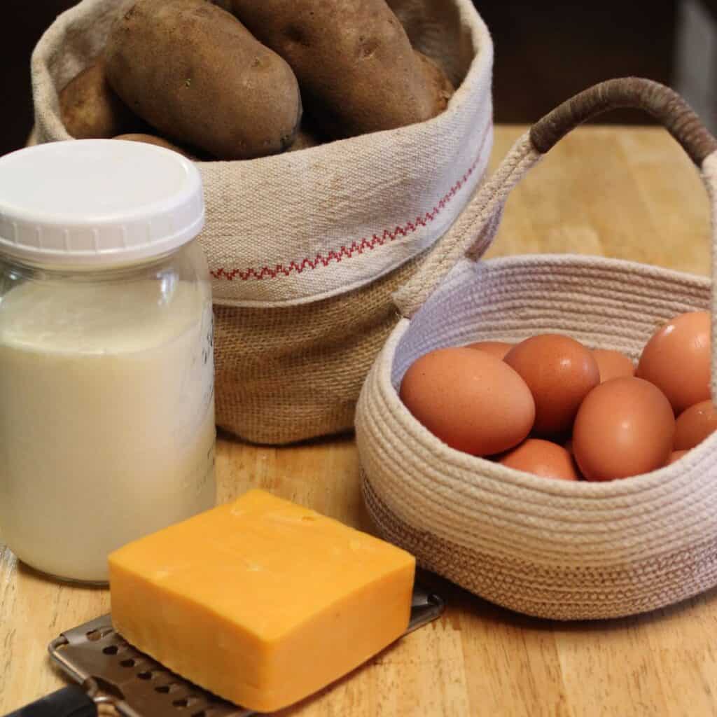 A jar of milk, a block of cheddar cheese on a grater, a basket of brown eggs, and a basket of potatoes are arranged on a wooden surface.