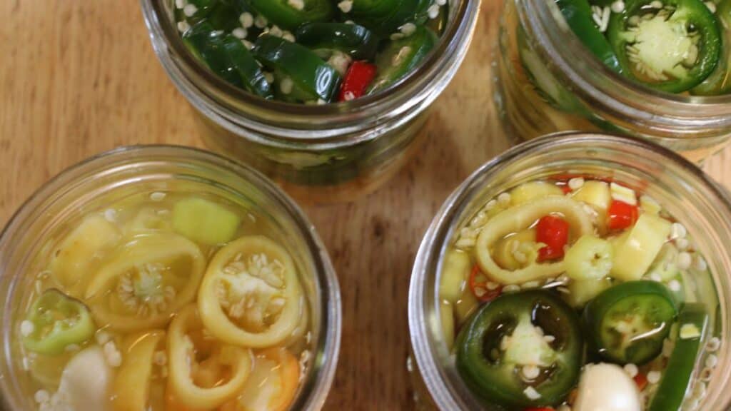 Four open glass jars filled with sliced fermented peppers in vibrant green, yellow, and red are arranged on a wooden surface. The peppers are submerged in liquid with visible seeds and garlic cloves.