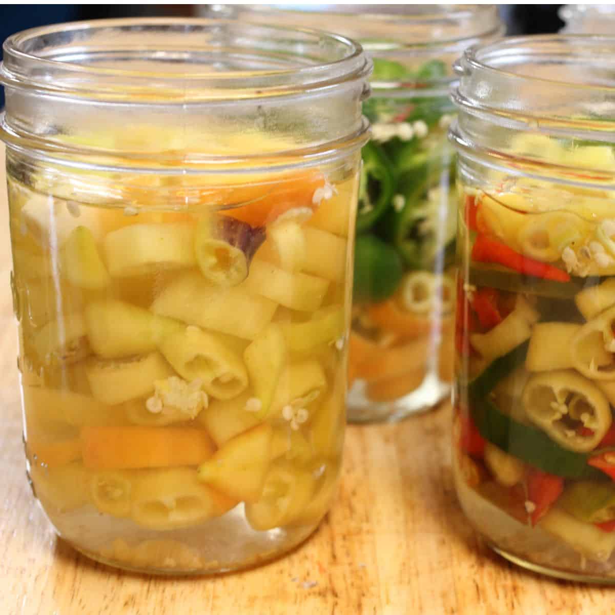 Three glass jars filled with sliced fermented peppers in a light brine sit on a wooden surface. The peppers, in yellow, green, and red hues, add a vibrant touch to the scene.