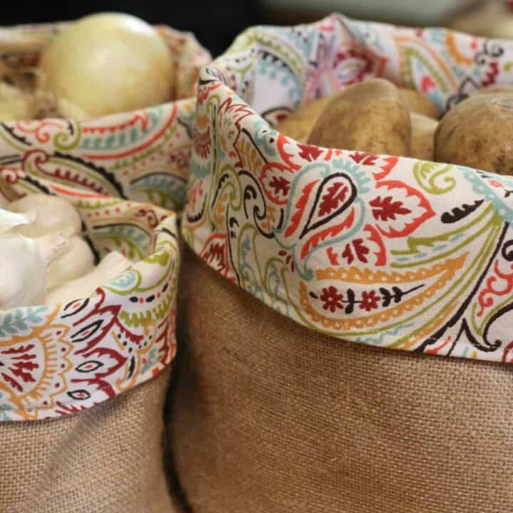 Two burlap bags with colorful, patterned fabric trim, one filled with onions and garlic, the other with potatoes. The bags are closely placed like a rustic produce basket, showcasing the fresh produce inside.