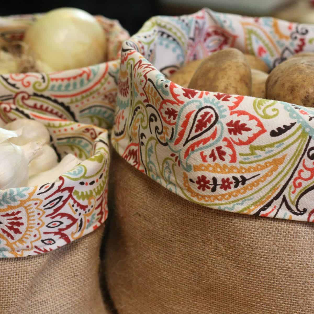 Two burlap bags with colorful, patterned fabric trim, one filled with onions and garlic, the other with potatoes. The bags are closely placed like a rustic produce basket, showcasing the fresh produce inside.