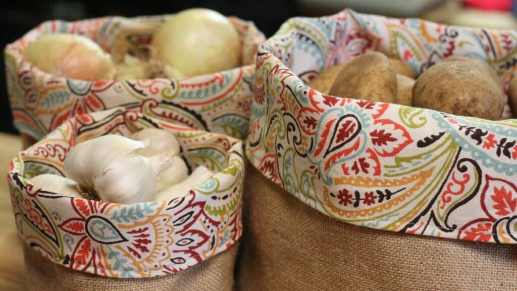 Three fabric-lined produce baskets made of burlap, filled with onions, garlic bulbs, and potatoes. The baskets feature a colorful, paisley-patterned trim and are arranged closely together on a countertop.