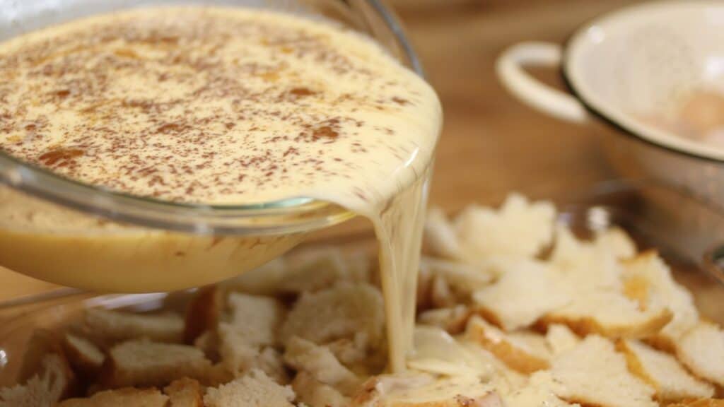 A glass bowl pours a creamy custard mixture sprinkled with spices over chopped bread pieces in a baking dish, preparing for baked french toast. A colander and eggs are blurred in the background.