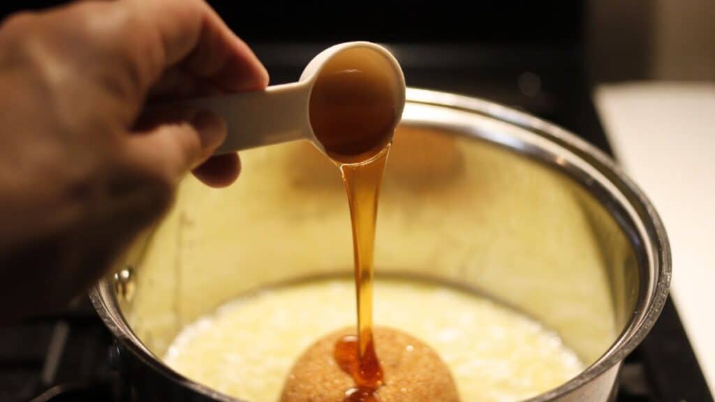 A hand holds a measuring spoon, pouring syrup or honey over a mound of brown sugar in a saucepan on a stovetop—perfect for making baked French toast.