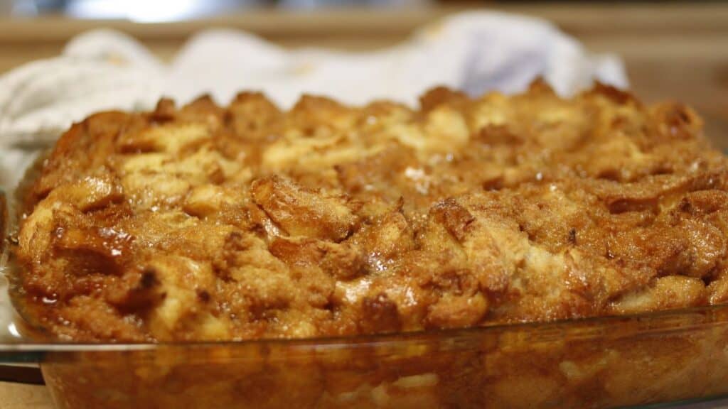 A close-up of a golden-brown baked French toast bread pudding in a glass baking dish, with a crispy, textured top and visible pieces of bread, set on a kitchen counter.
