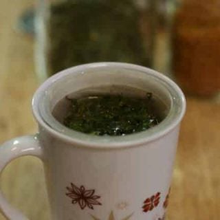 A white mug with floral designs filled with Moringa tea on a wooden surface. Loose green herbs are visible floating on top. The blurry background reveals jars of ingredients.