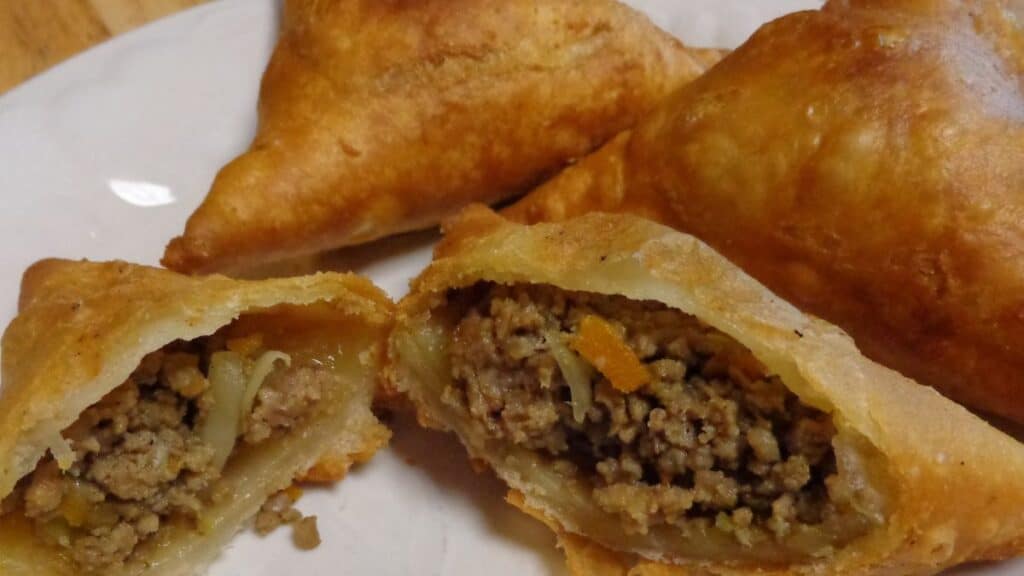 Golden-brown Kenyan Samosa pastries on a plate, one cut open to reveal a savory filling of ground meat and vegetables inside.