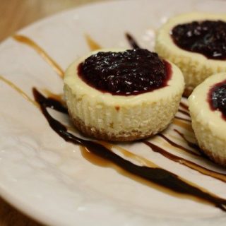 Three mini cheesecakes topped with a dark berry sauce grace a white plate, artistically drizzled with caramel and chocolate sauce.