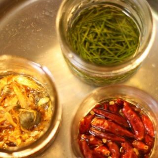 Three open glass jars simmer in hot water, each a haven for unique ingredients: one brimming with pine needles, another with fiery dried chilies, and the last with dried shrimp and onions. These exquisite combinations promise aromatic infused oils, captured beautifully from above.