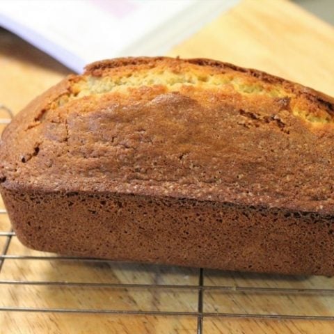 A freshly baked loaf of banana bread rests on a wire cooling rack on a wooden table. A book is open in the background.
