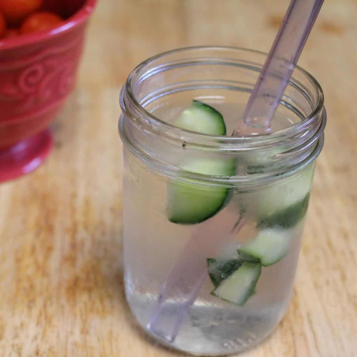 A mason jar filled with water, ice, and slices of cucumber—perfect if you freeze cucumbers for extra chill. In the background, a red bowl holds small orange objects, possibly cherry tomatoes, on a wooden surface.