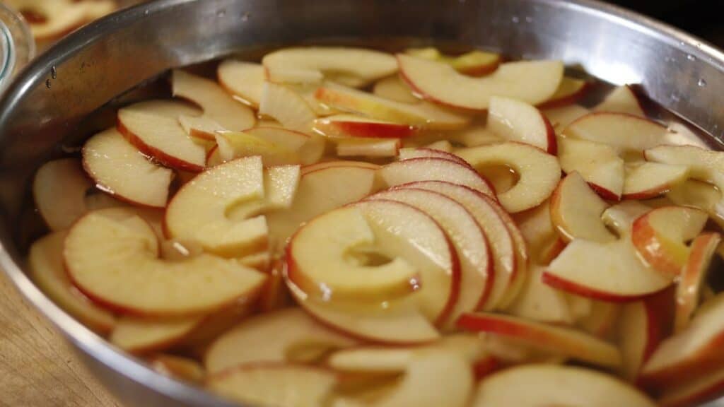 Sliced red apples soaking in a large stainless steel bowl filled with water, preparing them for dehydrating apples, with the slices partially submerged and evenly dispersed.