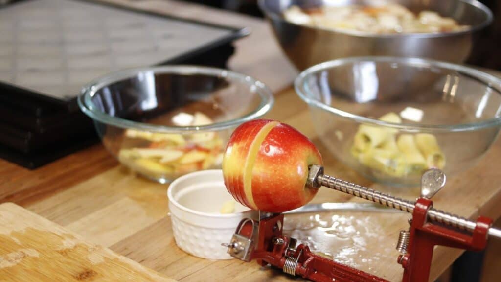 A red apple is being peeled and sliced with a hand-crank apple peeler on a wooden counter, perfect for dehydrating apples. Glass bowls with sliced apples and a metal bowl are in the background.