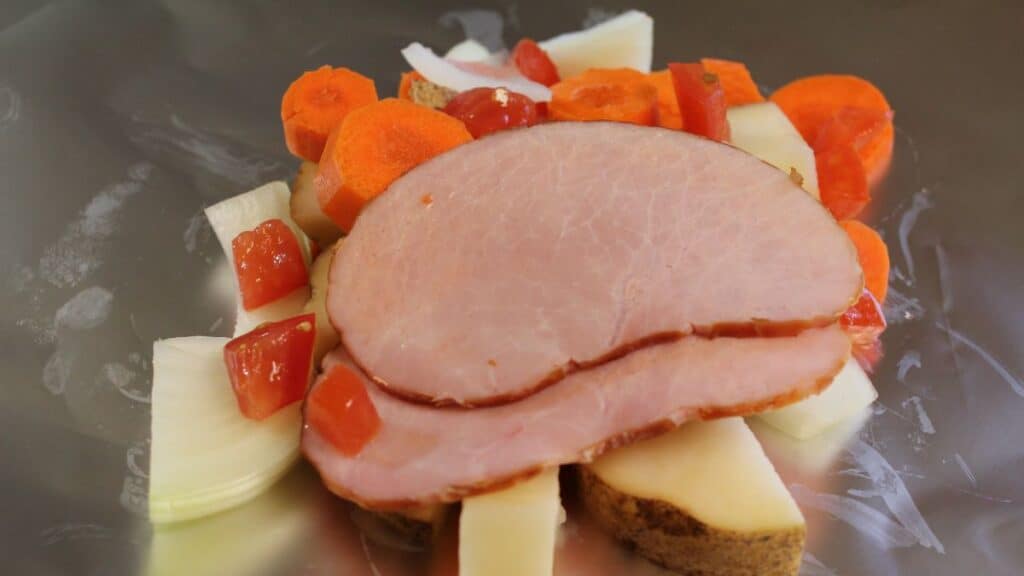 Two slices of ham are placed on chopped potatoes, carrots, onions, and diced tomatoes, arranged on a sheet of aluminum foil to create classic silver turtles.