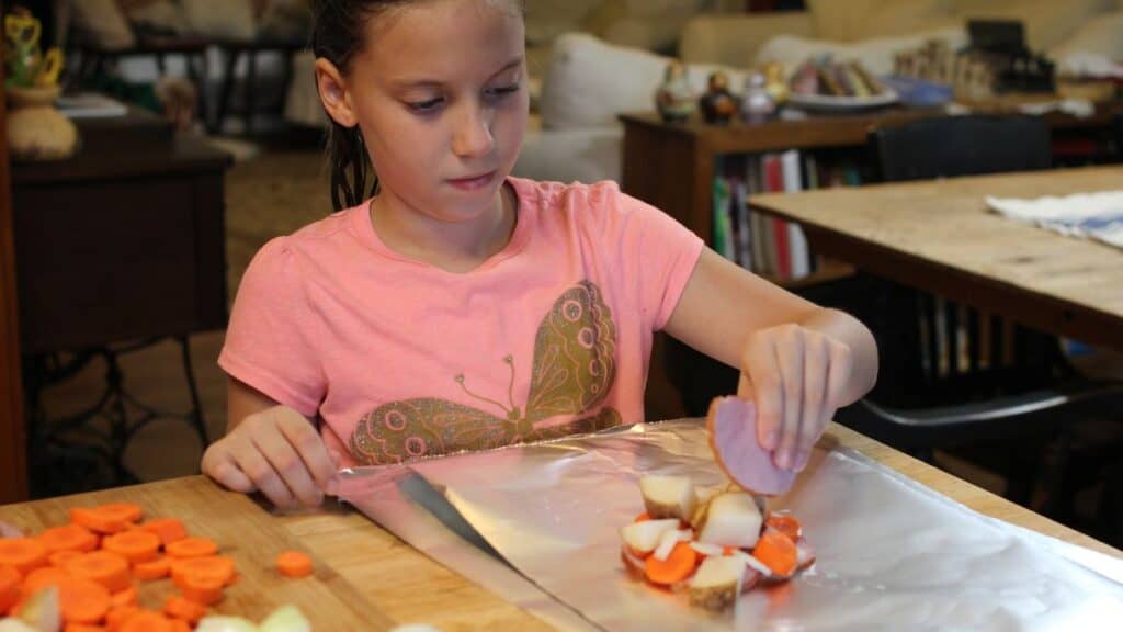 A girl in a pink shirt places sliced vegetables and a piece of ham on aluminum foil at a wooden table, preparing food. Sliced carrots, onions, and potatoes are nearby as she gets ready to make silver turtles.