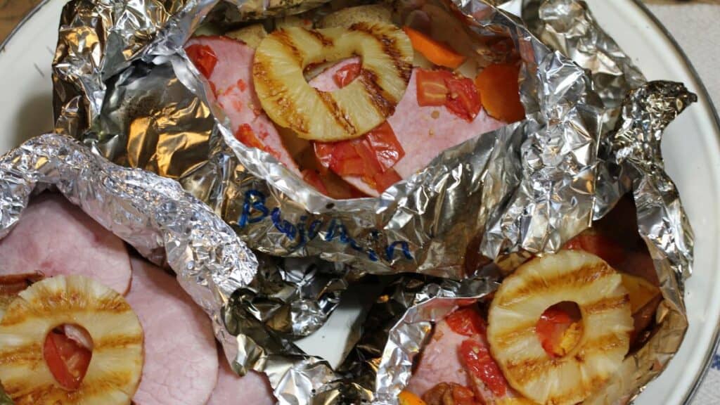 Slices of ham topped with pineapple rings and chopped red peppers are wrapped in foil to create delicious silver turtles, then placed on a white dish, ready for baking or serving.