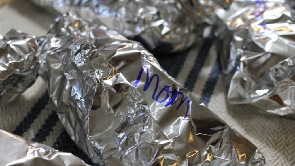 A foil-wrapped item labeled “Mom” in blue marker sits on a striped fabric surface, surrounded by other unmarked foil-wrapped packages resembling little silver turtles.