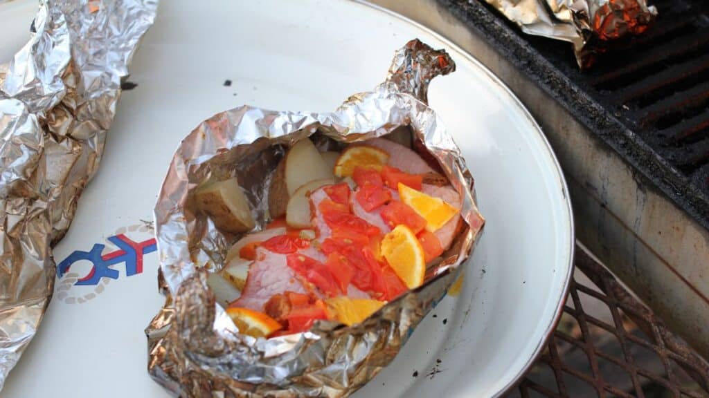 A foil packet on a plate, often called silver turtles, contains sliced potatoes, orange slices, and diced tomatoes, ready to be cooked or freshly cooked; a grill is visible nearby.