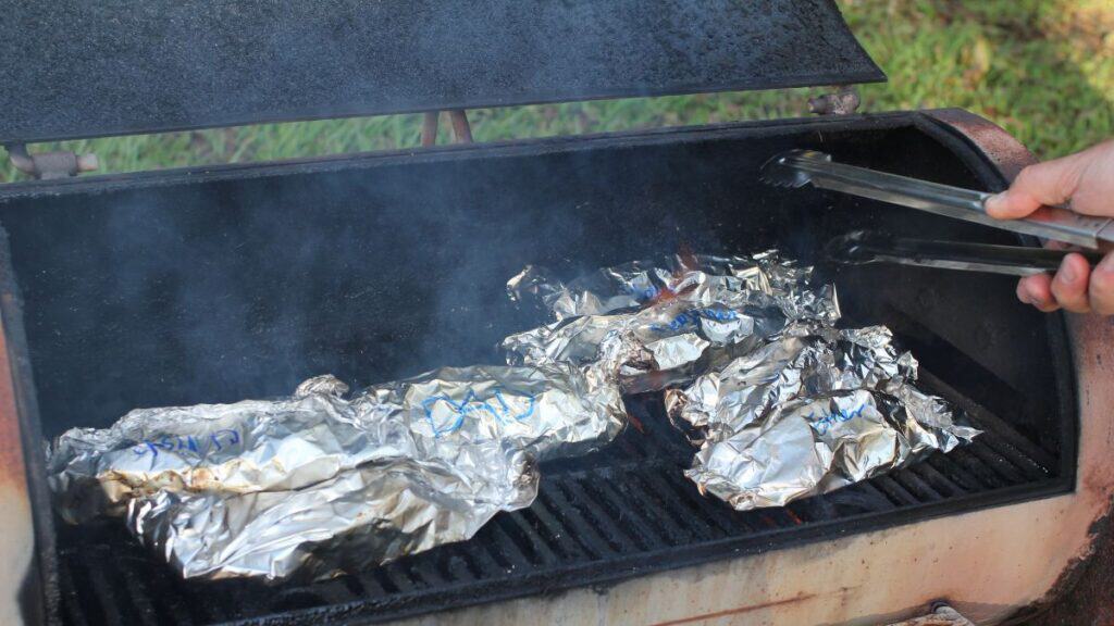 Several foil-wrapped packets, often called silver turtles, are being grilled on a smoky barbecue. A person holds tongs over the grill, ready to turn or remove the silver turtles. The background shows green grass.
