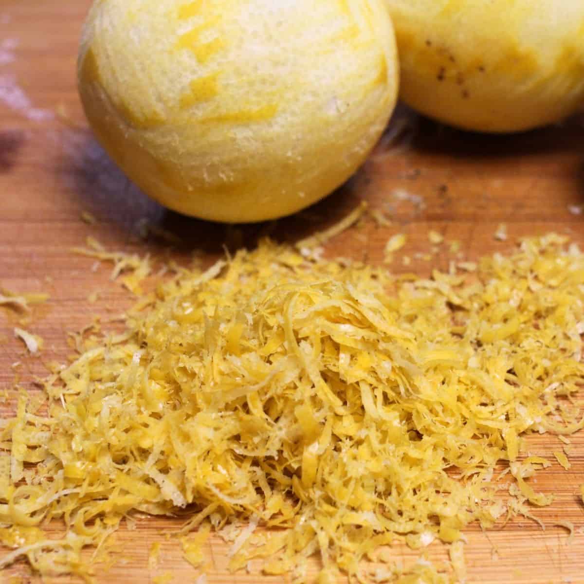 A pile of fresh lemon zest on a wooden surface with two partially peeled lemons in the background, perfect for adding a zing to recipes or to preserve Meyer lemons for later use.