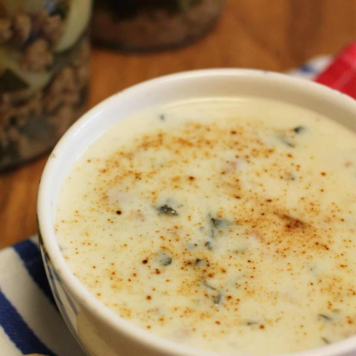 A bowl of creamy sausage potato kale soup with visible herbs, topped with a sprinkle of ground spices, sits on a striped cloth with blurred food jars in the background.